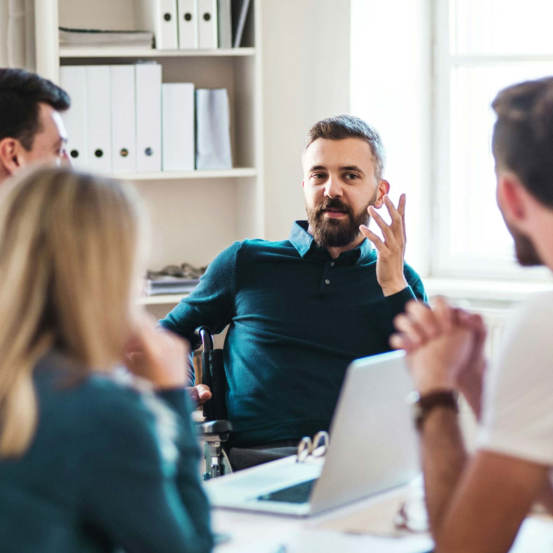 A bearded man wearing a polo shirt sitting in a wheelchair holding a meeting in an office with coworkers